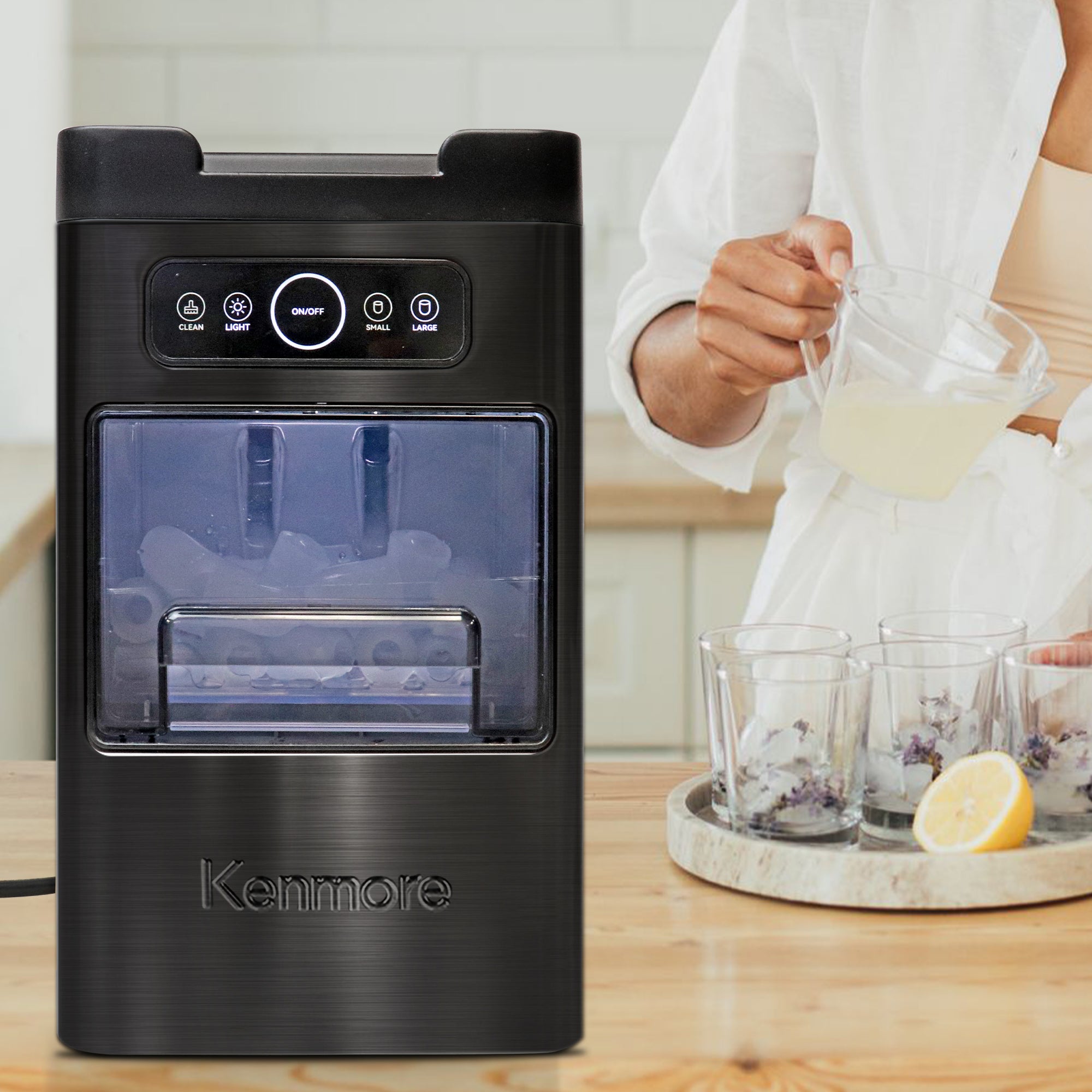 Kenmore automatic front-load ice maker on a light wooden countertop with a person pouring lemonade into glasses filled with ice in the background