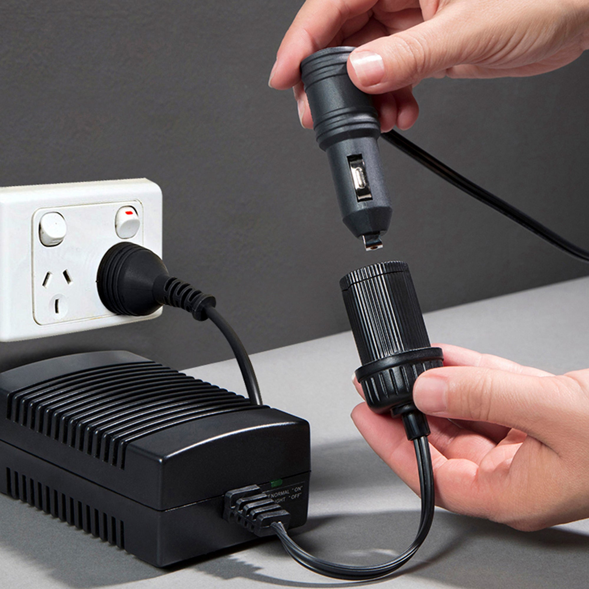 Koolatron AC to DC power adapter plugged into a white indoor AC receptacle on a dark gray wall with a person's hands inserting a 12V plug into the DC adapter end