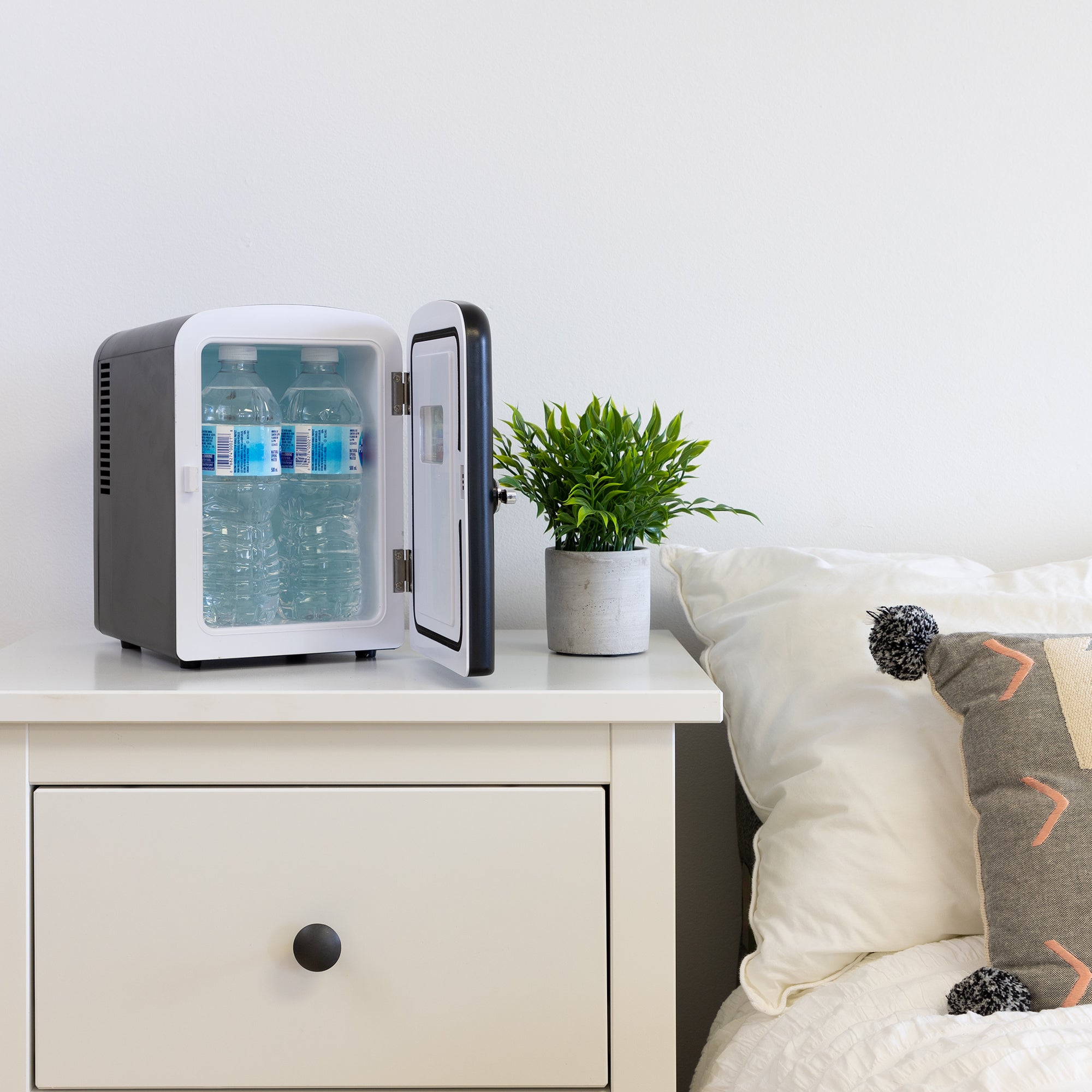 Lifestyle image of Koolatron retro 6 can mini fridge, open with 4 water bottles inside, with a plant in a white pot on its right, on a white bedside table beside a bed with white linens