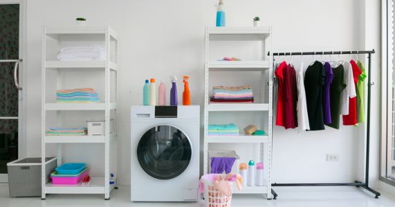 Picture of a laundry room with a front load washer in the middle of a white wall, white utility shelf units on either side, and a black metal clothing rack on the far right with several items of clothing on hangers.