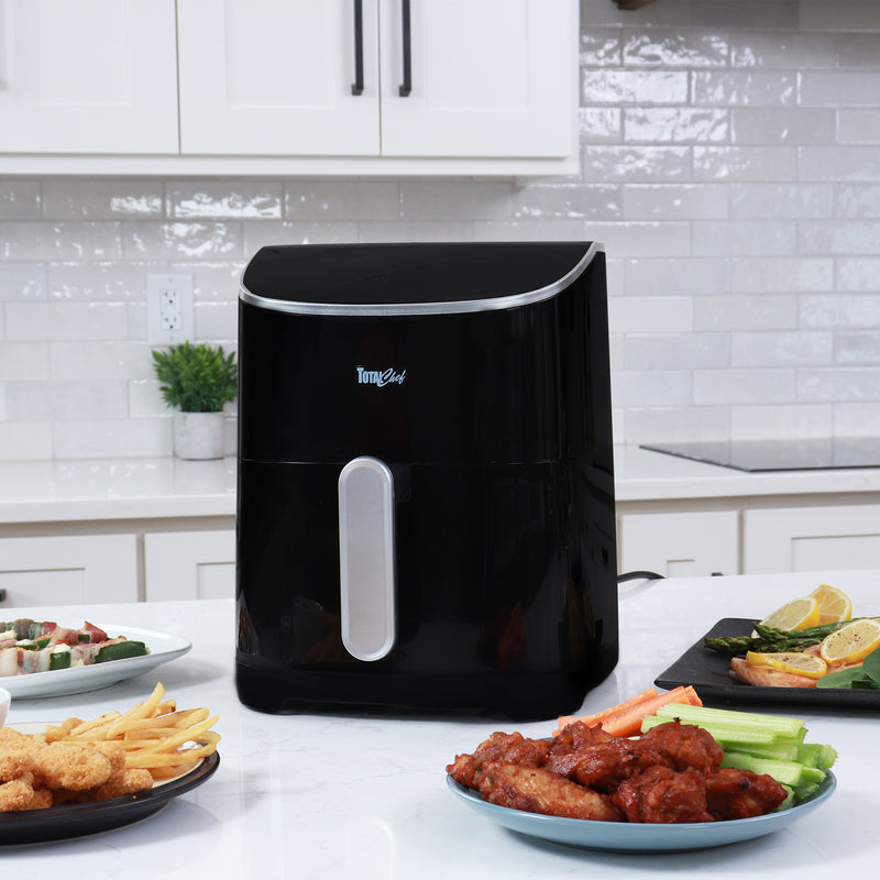Total Chef air fryer on a white kitchen counter with plates of cooked food arranged around it and white cupboards and white tile backsplash in the background.