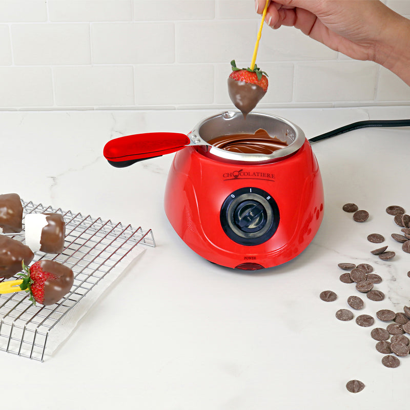 Lifestyle image of a hand dipping a strawberry in melted chocolate in the chocolatiere on a white countertop. There is a cooling rack to the left with dipped strawberries and marshmallows on it and unmelted chocolate discs scattered on the counter to the right