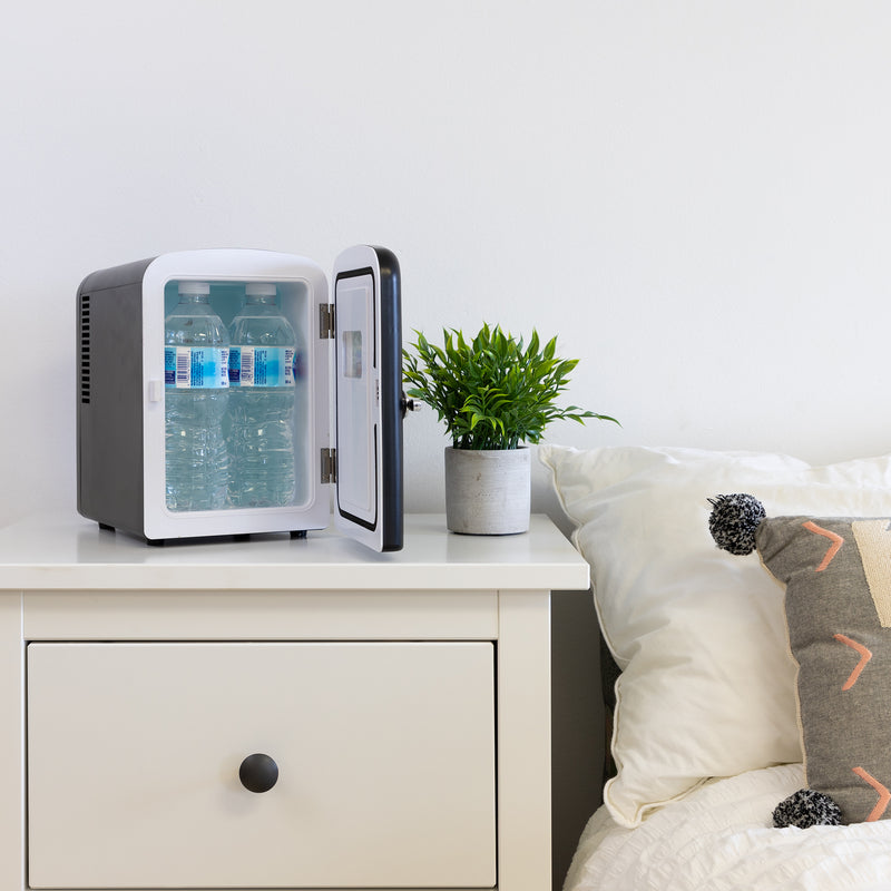 Lifestyle image of Koolatron retro 6 can mini fridge, open with 4 water bottles inside, with a plant in a white pot on its right, on a white bedside table beside a bed with white linens 