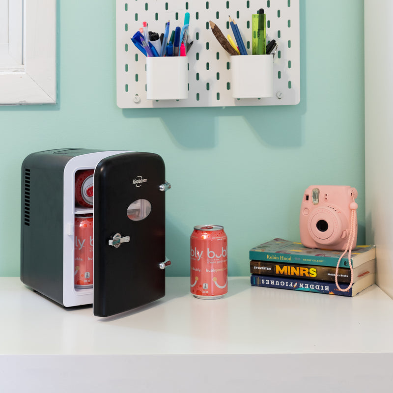 Lifestyle image of Koolatron retro 6 can mini fridge, partly open with cans inside, on a white desktop with an aqua wall behind. There is a can of soda, a stack of books, and a pink camera to the right of the fridge and two cups of pens and pencils attached to the wall above